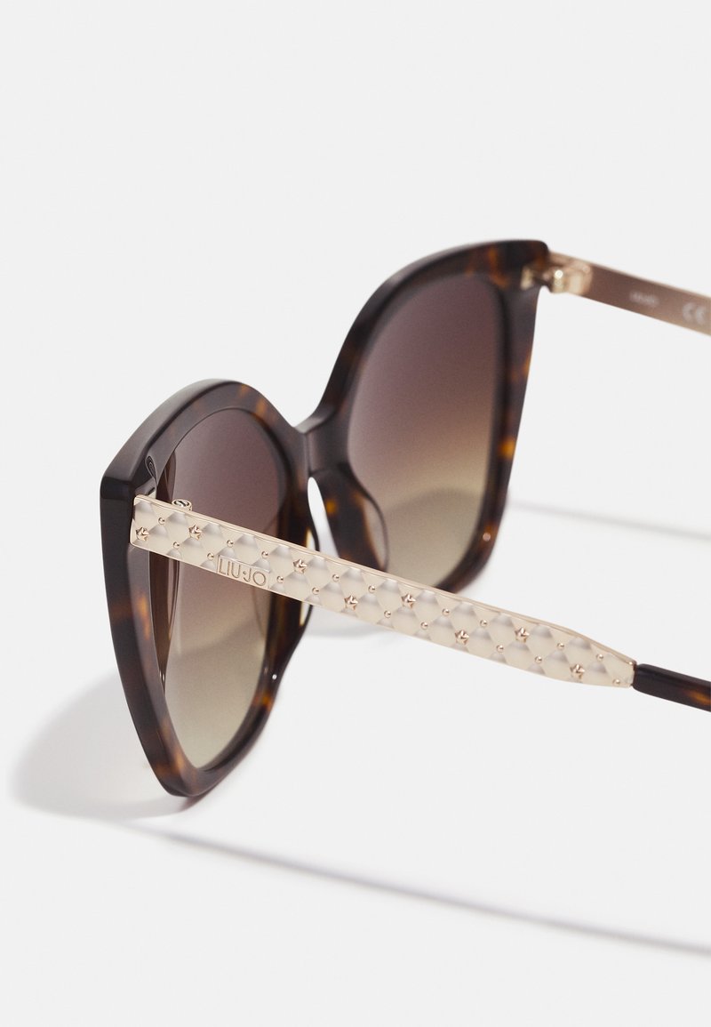 Sonnenbrille aus Schildpatt mit Katzenaugenform, getönten Gläsern und beigen strukturierten Bügeln mit metallischen Akzenten und Logodetail.