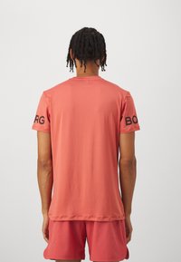 T-shirt corail à manches courtes en tissu lisse, avec un col rond et des lettres noires sur la manche gauche.