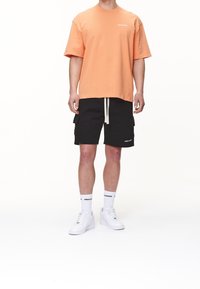 T-shirt arancione a maniche corte con piccolo logo bianco, abbinato a pantaloni cargo neri con tasche laterali e stringa beige. Sneakers bianche e calze.