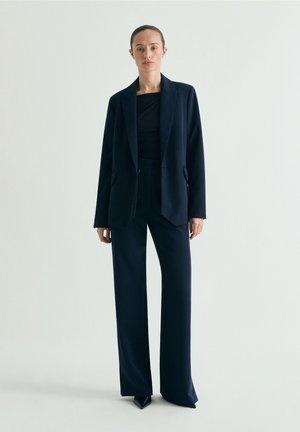 Mujer de pie vestida con un traje azul marino oscuro con una blazer larga, pantalones de pierna ancha y zapatos negros puntiagudos contra un fondo claro y liso.