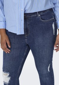Jeans de mezclilla azul oscuro con desgastes y rasgaduras en el muslo y la rodilla. Presenta un corte ajustado y un estilo clásico de cinco bolsillos. Combinados con una camisa a rayas.