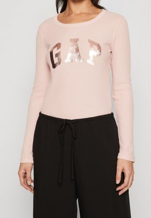Femme portant un haut rose clair à manches longues avec un lettrage brillant GAP et un pantalon noir ample avec cordon.