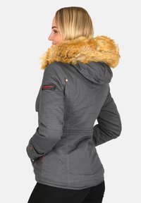 Woman wearing a gray winter jacket with a large brown fur-trimmed hood, standing in profile with hands in pockets.