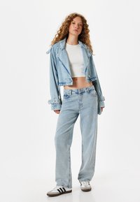 Chaqueta de mezclilla cortada en un azul claro con un gran cuello y puños con cinturón, combinada con pantalones vaqueros de pernera ancha en azul claro y zapatillas blancas.