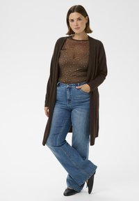 Brauner Cardigan über einem transparenten braunen Oberteil mit silbernen Punkten, kombiniert mit blauen, hoch taillierten Schlagjeans und schwarzen Schuhen. Einfaches Design und Texturen.
