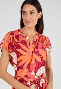 Robe florale avec un motif vibrant rouge et rose, manches courtes et décolleté en V avec une taille nouée. Fabriquée en tissu léger.