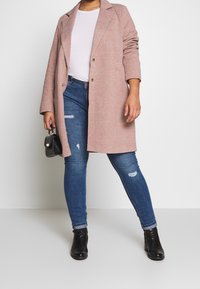 Vaaleanpunainen pitkä takki, jossa on tekstuuri, läppäkaulus ja kaksi etupainiketta, yhdistettynä revittyihin sinisiin farkkuihin ja mustiin nilkkureihin.