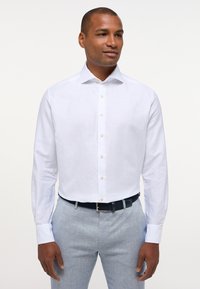 Camicia bianca a maniche lunghe con colletto classico, dotata di bottoni beige chiaro, abbinata a pantaloni grigi chiari e una cintura intrecciata scura.