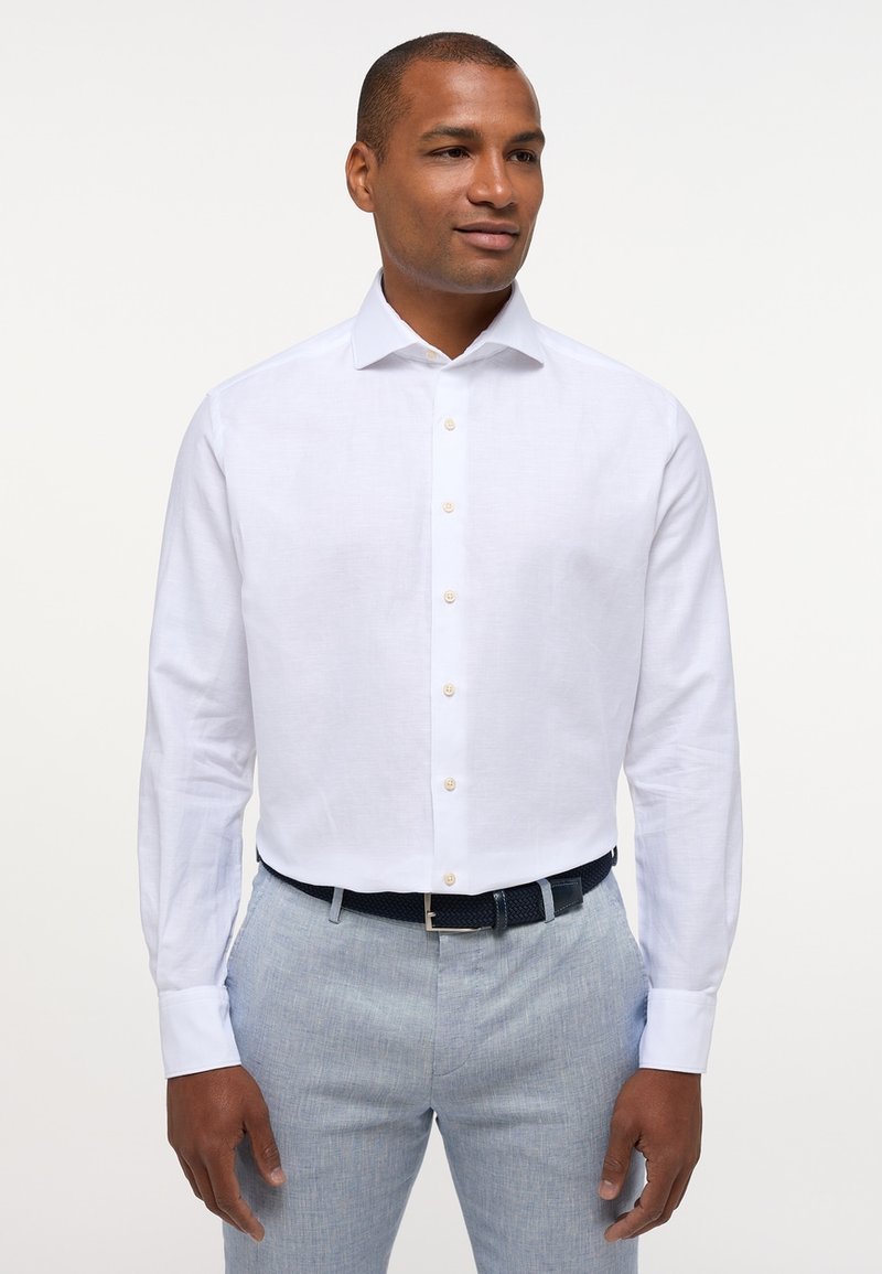 Camicia bianca a maniche lunghe con colletto classico, dotata di bottoni beige chiaro, abbinata a pantaloni grigi chiari e una cintura intrecciata scura.