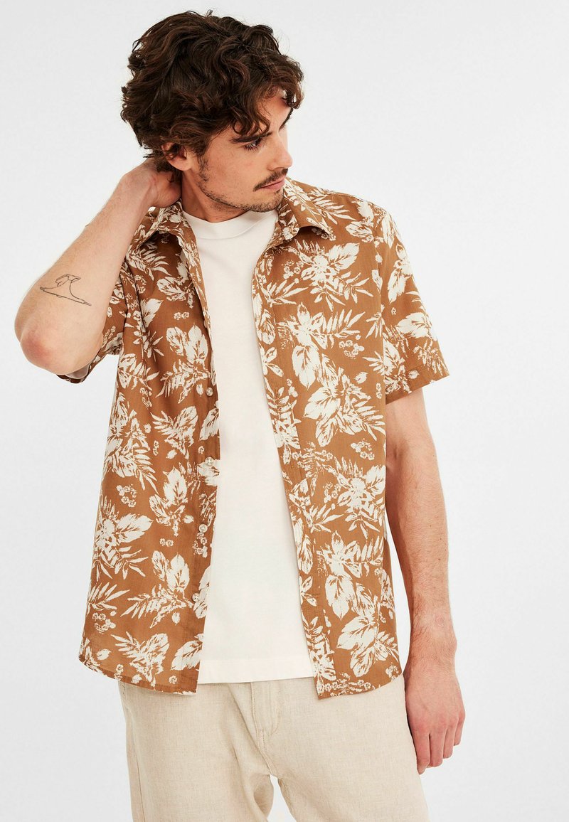 Koszula z krótkim rękawem w brązowe kwiaty z białymi wzorami liści, zapinana na guziki, z lekkiego materiału, noszona na białym T-shircie, w luźnym fasonie.