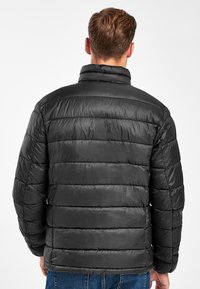 Giacca nera imbottita con colletto alto, caratterizzata da cuciture orizzontali e una texture liscia. La vista posteriore mostra un design a taglio dritto.