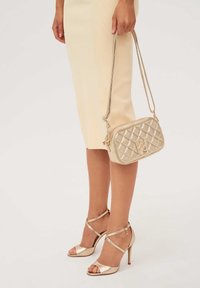 Kankaan kullanainen käsilaukku, jossa on vetoketjukiinnityksen ja hoikka kantohihna, yhdistettynä kiiltäviin kultaisiin nauhakorkokenkiin, vaaleaa taustaa vasten.