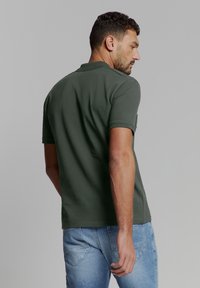 Donkergroene polo met korte mouwen, gemaakt van zacht materiaal. Heeft een klassieke kraag, subtiele stiksels en een casual pasvorm. Gecombineerd met lichtblauwe jeans.