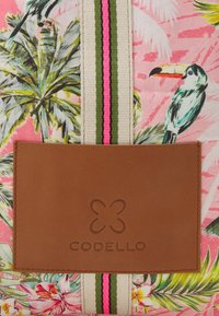 Torba na ramię z różowym tropikalnym nadrukiem przedstawiającym liście palmowe i tukany, brązowa skórzana łatka z tłoczonym logo oraz paski tkaniny jako dodatki.