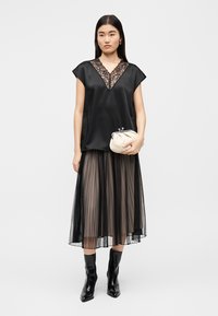 Schwarzes Satintop mit Spitzen-Ausschnitt, kombiniert mit einem plissierten Rock in Schwarz und Taupe. Das Model hält eine weiße Clutch und trägt schwarze Stiefeletten.