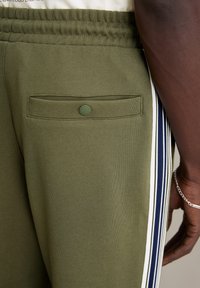 Olivengroene sweatpants met een achterzak met drukknoopsluiting, contrasterende witte en marineblauwe zijstrepen, en een gladde textuur.