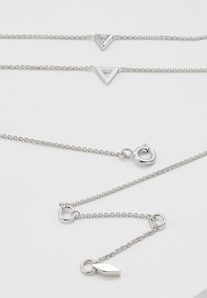 Deux colliers en chaîne argentée avec des pendentifs triangulaires, l'un plein et l'autre creux, avec un fermoir portant une petite étiquette Fossil sur un fond blanc.