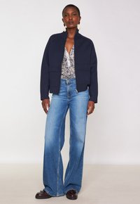Donna che indossa una giacca zip blu navy sopra una blouse con motivo, jeans a gamba larga blu, mocassini marroni scuri e grandi orecchini a goccia, in piedi contro uno sfondo semplice.