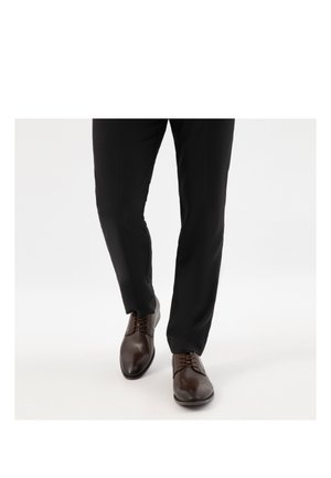 Mann trägt schwarze Hose und polierte braune Lederschuhe, geht auf einem schlichten weißen Hintergrund.