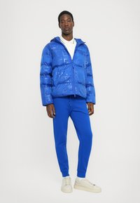 Chaqueta acolchada azul con capucha, textura brillante, cierre de cremallera y diseño estampado; combinada con pantalones de chándal azul sólido y zapatillas blancas.