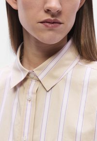 Beige Hemd mit vertikalen lila Streifen, Knopfleiste und Knöpfen vorne. Der Stoff wirkt glatt mit einem dezenten Glanz.