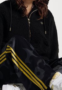 Sudadera con cremallera de forro polar negro con capucha, herrajes dorados y logotipo bordado. Pantalones negros con patrón y rayas amarillas y zapatillas blancas.