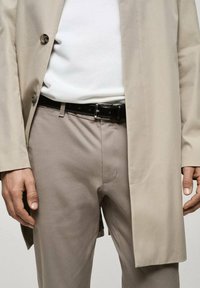 Béžový kabát s velkými knoflíky, pod ním bílá košile, černý kožený pásek a světle béžové kalhoty s hladkou texturou a na míru střižené.