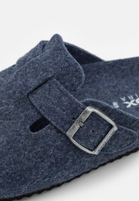 Zoccoli in feltro blu scuro con una sola cinghia e una fibbia metallica, caratterizzati da una superficie di tessuto testurizzato e un design del plantare anatomico.