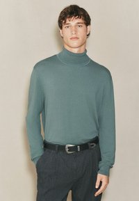 Pulover cu guler înalt din material tricotat de un verde moale, cu mâneci lungi și un guler ajustat, asortat cu pantaloni negri texturați, croiți, și o curea neagră.