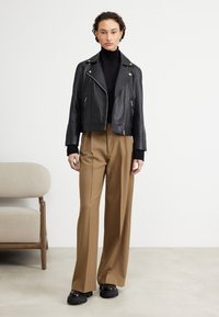 Schwarze Leder-Motorradjacke in verkürzter Form, schwarzes Oberteil mit hohem Kragen, braune weit geschnittene Hose und klobige schwarze Plateauschuhe. Minimalistisches Design.