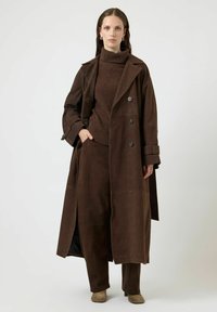 Cappotto lungo in camoscio marrone con design doppiopetto, ampi revers e tasche laterali, abbinato a un dolcevita e pantaloni coordinati.