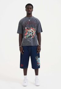 Grafinen harmaa t-paita, jossa on pääkalloasu, yhdistetty tummiin farkkushortseihin, joissa on värikäs brodeeraus, ja valkoisiin lenkkareihin.