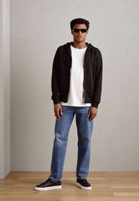 Schwarze Zip-Up-Hoodie, weiße T-Shirt, hellblaue Jeans und schwarze Sneakers. Das Model trägt dunkle Sonnenbrillen und steht auf einem Holzboden vor einer grauen Wand.
