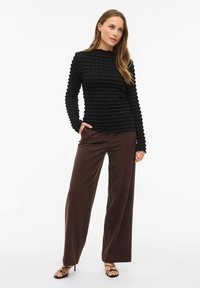 Maglione nero con una scollatura ondulata, maniche lunghe e un design aderente, abbinato a pantaloni larghi marroni e tacchi neri con cinturini.