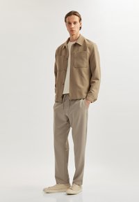 Tan skjortejakke med to store brystlommer, b�året over en lys t-shirt. Kombineret med beige bukser med snøre og lyse sneakers.