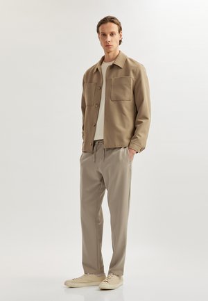 Beige shirtjacket met twee grote borstzakken, gedragen over een lichtgekleurde t-shirt. Gecombineerd met beige broek met trekkoord en lichte sneakers.