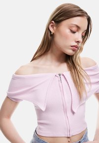 Lyserød ribbet cropped top med off-the-shoulder ærmer og lynlåsdetalje foran. Blød tekstur, tætsiddende design og minimalistisk stil.