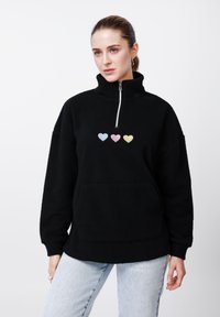 Μαύρο φλις πουλόβερ με ψηλό γιακά, διαθέτει φερμουάρ τετάρτου μήκους και τρεις πολύχρωμες καρδιές (μπλε, ροζ, κίτρινη) στο στήθος.