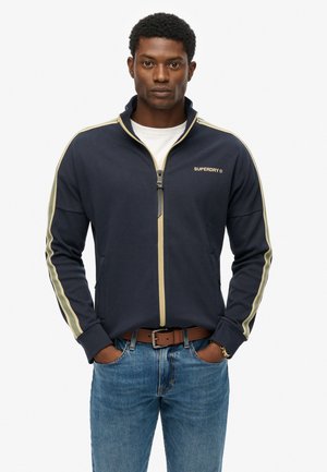 Marineblauwe zip-up jas met lichtgroene strepen op de mouwen, gemaakt van zachte stof. Kenmerken zijn een hoge kraag en voorzakken.