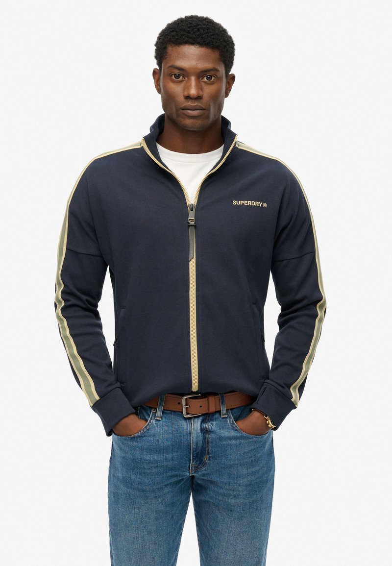 Marineblauwe jas met rits en crèmekleurige strepen aan de zijkant, gemaakt van zachte stof. Heeft een hoge kraag, een gouden logo en zakken aan de voorkant.