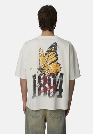 Maglietta oversize color crema con una grande grafica di una farfalla gialla e nera e il numero "1894" in un carattere nero consumato sulla schiena.