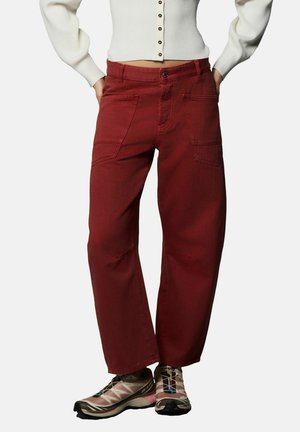 Piros színű, pamut anyagú cargo nadrág, laza szabással, nagy első zsebekkel és gombos rögzítéssel. Bézs sportcipővel viselve.