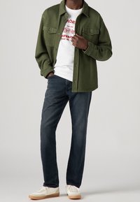 Grön knapp-up jacka över en vit grafisk t-shirt, mörka jeans och vita sneakers med bruna sulor. Outfiten har en avslappnad stil.