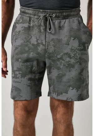 Short à cordon avec motif camouflage gris et noir sur un homme les mains dans les poches et une chemise foncée rentrée.