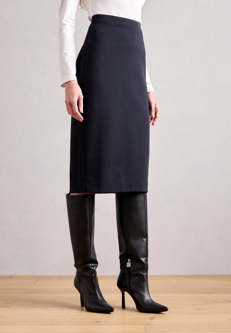 Femme portant une jupe crayon noire longueur genou, un haut blanc à manches longues et des bottes noires à talons hauts jusqu'aux genoux, debout sur un sol en bois.