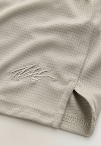 T-shirt de sport gris clair en tissu texturé avec un motif à grille, présentant un logo brodé dans le coin inférieur et des ourlets cousus.