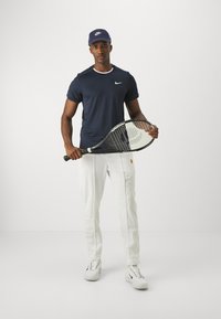 Marineblå kortermet Nike-skjorte med hvite detaljer, hvite treningsbukser, holder en tennisracket, iført en marineblå Nike-cap og hvite joggesko.