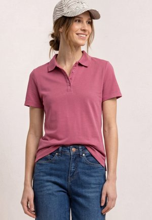 Vrouw glimlacht, draagt een mauve poloshirt, blauwe spijkerbroek en een pet met zebrapatroon, staand tegen een eenvoudige lichte achtergrond.