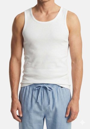 Mies, jolla on valkoinen hihaton toppi ja vaaleansiniset kiristysnauhalla varustetut housut, seisoo yksiväristä taustaa vasten.