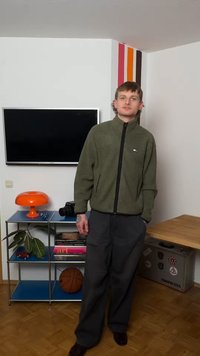 Young man in green jacket and dark pants stands in room with wall-mounted TV, orange lamp, basketball, camera, and colorful wall stripes.